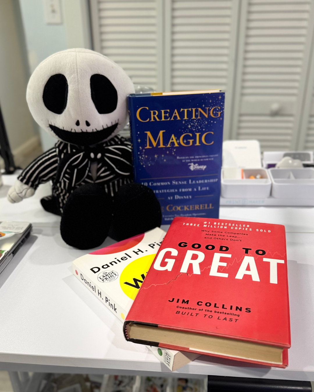 GPT-4o
The image features a plush toy resembling Jack Skellington from “The Nightmare Before Christmas” sitting on a table. Next to it are three books: "Creating Magic" by Lee Cockerell, "When" by Daniel H. Pink, and "Good to Great" by Jim Collins. White storage drawers are visible in the background.